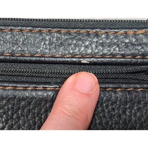 May include: Close-up of a black leather zippered wallet with brown stitching. The zipper is visible and the leather has a textured grain.