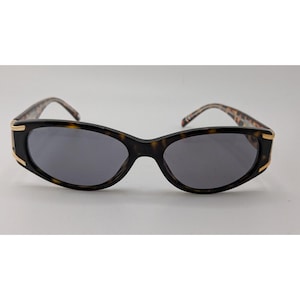 May include: Oval sunglasses featuring a dark tortoiseshell frame with gold-tone metal accents at the outer hinges. The lenses are dark grey and provide full coverage. The temples display a matching tortoiseshell pattern with a smooth finish.