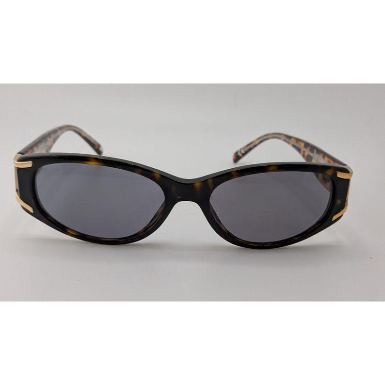May include: Oval sunglasses featuring a dark tortoiseshell frame with gold-tone metal accents at the outer hinges. The lenses are dark grey and provide full coverage. The temples display a matching tortoiseshell pattern with a smooth finish.