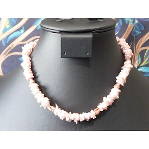 May include: A light pink necklace made of small, irregularly shaped pieces. The necklace is displayed on a black mannequin. The background features a dark blue fabric with a floral pattern. The necklace is a delicate piece of jewelry.