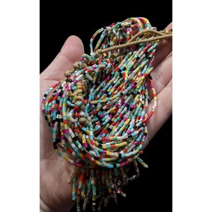 May include: A colorful beaded necklace with a multitude of small, rectangular beads in various colors, including red, yellow, blue, and black. The beads are strung together, creating a cascading effect. The necklace is held in a hand against a black background.