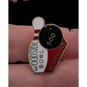 May include: A gold-toned metal enamel pin featuring a white bowling pin with red stripes and the text Woodside Bowl, alongside a black bowling ball with three finger holes and the number 500, with the text Blumenfeld printed in red.