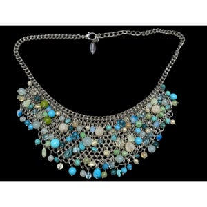 Peut inclure: Un collier argenté avec une chaîne et une cascade de perles colorées. Les perles sont dans des tons de bleu, vert et blanc, créant un design vibrant et accrocheur. Le collier est posé sur un fond noir.
