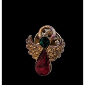 May include: A vintage angel pin with a gold-tone finish, featuring a green gemstone at the center and a teardrop-shaped red gemstone. The angel has detailed wings and is set against a black background.
