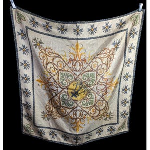 May include: Square scarf with a cream background and intricate patterns in brown, green, and yellow. The design includes a central emblem and a border of cross-like shapes. The scarf is hanging against a black background.