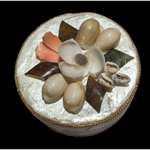 May include: A round, white, decorative box with a gold trim. The lid is decorated with seashells arranged in a flower shape. The flower has a white center and pink petals.