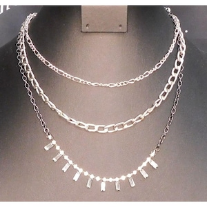 May include: A three-strand necklace set displayed on a black mannequin. The top chain is a delicate silver chain, the middle chain is a thicker silver chain, and the bottom chain features small rectangular crystals.