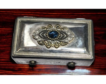 May include: A silver rectangular box with a decorative gold and blue design on the lid. The box has a beaded border and two small metal feet.