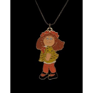 May include: A gold necklace with a charm of a red-haired girl wearing a yellow dress with white polka dots. The girl has a red flower crown and blue eyes.
