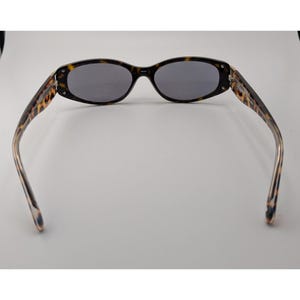 May include: Oval sunglasses featuring a brown and black tortoiseshell pattern frame with dark gray lenses. The temples extend outward from the frame, displaying the same tortoiseshell pattern, and the bridge connects the two oval-shaped lenses.