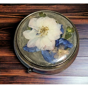 Peut inclure: Une boîte à bijoux ronde en laiton avec un couvercle en verre orné de fleurs pressées. Les fleurs sont blanches, bleues et vertes. La boîte a un couvercle à charnière et un petit bouton décoratif.