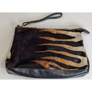May include: A rectangular clutch bag with a black leather base and wrist strap. The main body features a natural animal print pattern in shades of brown, black, and tan. The zipper is gold-toned.
