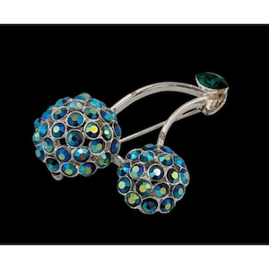 May include: A silver-toned brooch in the shape of cherries. The cherries are made of blue and green iridescent crystals. A small green crystal leaf accents the stem. The brooch is set against a black background.