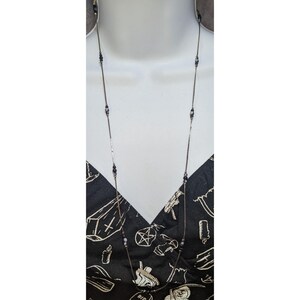 May include: A long, thin silver-tone chain necklace featuring small, evenly spaced black and grey beads. The necklace is displayed against a black fabric garment printed with white line drawings of coffins, candles, and pentagram symbols.