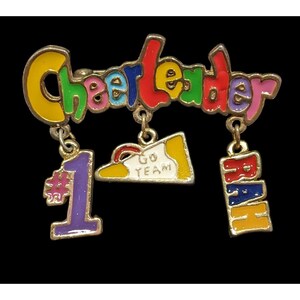 May include: A colorful cheerleader pin with the word "Cheerleader" in rainbow colors. The pin has three dangling charms: a number 1, a shoe with "Go Team" text, and the word "RAH".