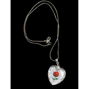 Pode incluir: Um colar de corrente de prata com um pingente em forma de coração que contém uma rosa vermelha dentro. O pingente é feito de vidro transparente.