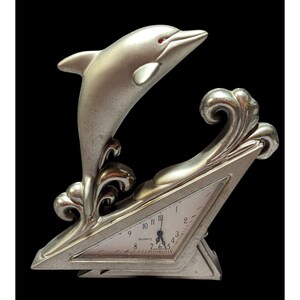 May include: A silver-toned clock shaped like a triangle with a dolphin jumping out of the top. The clock face is white with black numbers and hands. The clock says "Quartz" on the face.