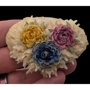 May include: An oval-shaped brooch featuring a cluster of three dimensional flowers made from small shells. The flowers are colored in yellow, pink, and blue, surrounded by cream-colored shell petals and small green shell leaves on a textured base.