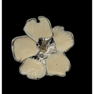 Puede incluir: Broche con forma de flor con acabado esmaltado color crema y ribete metálico plateado. El centro del broche presenta un adorno dorado y una pequeña piedra gris. El broche está sobre un fondo negro.