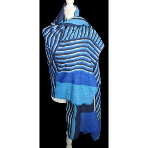 Può includere: Sciarpa a righe blu e bianche con un mix di sezioni a tinta unita e a righe. La sciarpa è drappeggiata su un manichino bianco, mostrando il suo design e la sua consistenza. La tavolozza dei colori della sciarpa comprende sfumature di blu e bianco.