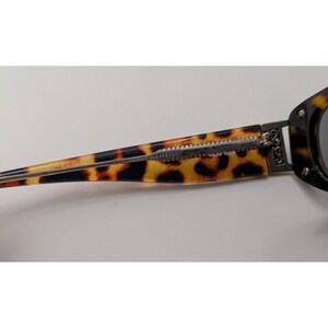 May include: Tortoiseshell patterned sunglasses temple arm featuring a silver-toned metal hinge and decorative side detail. The text "Haute Spots" is printed in small white lettering on the inner side of the brown and black patterned arm.