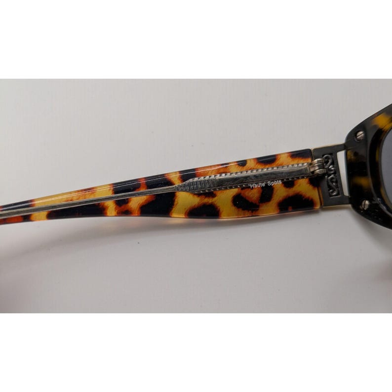 May include: Tortoiseshell patterned sunglasses temple arm featuring a silver-toned metal hinge and decorative side detail. The text "Haute Spots" is printed in small white lettering on the inner side of the brown and black patterned arm.