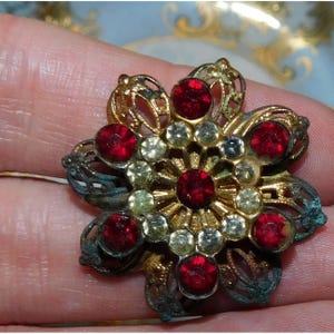 May include: Vintage floral brooch with a gold-tone filigree design. The brooch features red and clear rhinestones arranged in a flower shape. The brooch has a tarnished, aged appearance.