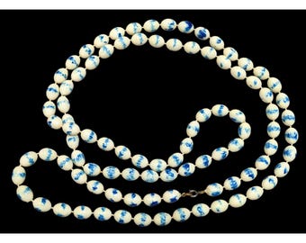 Puede incluir: Un collar largo con cuentas ovaladas en crema y azul. Las cuentas están ensartadas, creando un efecto en capas. El collar se muestra sobre un fondo negro, resaltando los colores y el diseño.