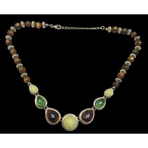 Puede incluir: Un collar con una cadena plateada y varias piedras preciosas. Presenta piedras en forma de lágrima marrones, verdes y amarillas, así como cuentas redondas marrones. El collar está sobre un fondo negro.