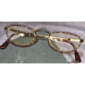 Puede incluir: Gafas de forma ovalada con montura dorada y estampado carey en los bordes y patillas. Las lentes son transparentes y las gafas descansan sobre una superficie gris. Las gafas tienen una estética clásica y vintage.
