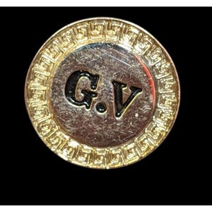 May include: A round, gold-toned button with a Greek key border. The center features the black letters "G.V." on a silver background. The button is set against a black backdrop.