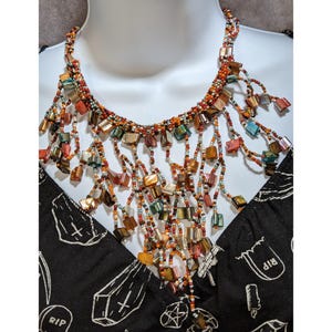 Op de afbeelding: Een meerstrengs kralenketting met een levendige reeks oranje, bruine en blauwgroene kraaltjes, afgewisseld met gepolijste, vierkante schelpaccenten. Het ontwerp bevat talloze bungelende strengen die een gelaagd franje-effect creëren.