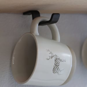 May include: A cream-colored ceramic mug with a deer design, hanging from a black metal hook. The mug has a rounded shape and a handle. The deer design appears to be made of text.