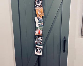 May include: A dark sage green sliding barn door with black hardware and a vertical hanging display of seven rectangular holiday photo cards. The cards feature various family portraits and the text Merry Christmas and Happiest Holidays.