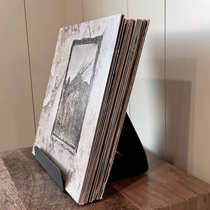 May include: A stack of vinyl records in a black metal display stand. The album cover features a sepia-toned image within a frame. The records are displayed on a wooden surface against a white wall.