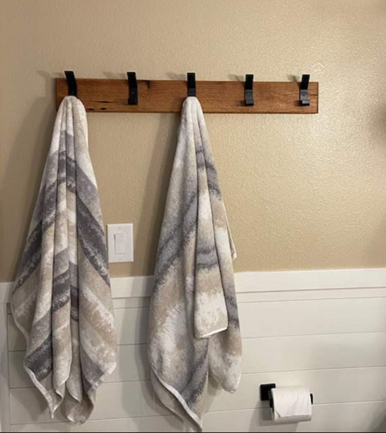 May include: A wooden towel rack with five black metal hooks. Two towels are hanging on the hooks. The towels are a light gray and white pattern.