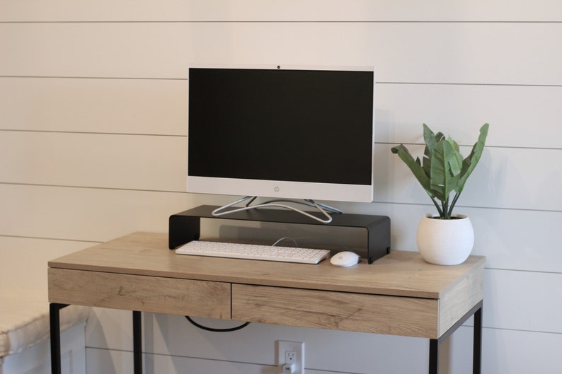 Puede incluir: Un monitor de ordenador blanco se encuentra sobre un soporte de metal negro en un escritorio de madera marr&oacute;n claro con un teclado y un rat&oacute;n blancos. Una planta verde en maceta se encuentra en el escritorio a la derecha del monitor.