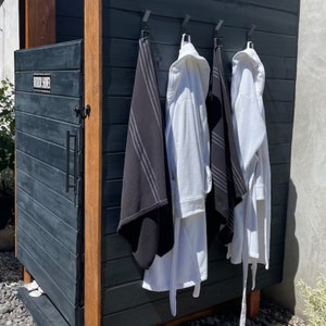 May include: A black outdoor shower with a wooden frame and a black door. Three white bathrobes and a black towel are hanging on hooks on the wall.