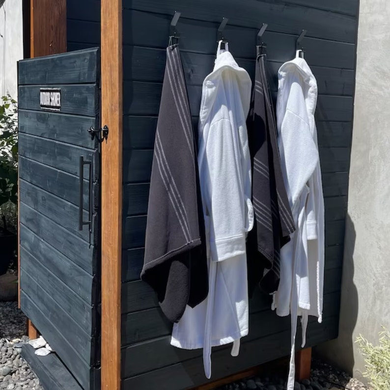 May include: A black outdoor shower with a wooden frame and a black door. Three white bathrobes and a black towel are hanging on hooks on the wall.