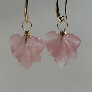 May include: A pair of earrings featuring carved pink leaf-shaped stones. The leaves are suspended from gold-colored hooks with gold wire accents. The earrings are displayed against a neutral background.