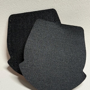 May include: Two black fabric patches with a textured surface. The patches are in a shield shape with rounded corners and a notched bottom. One patch is slightly darker than the other.