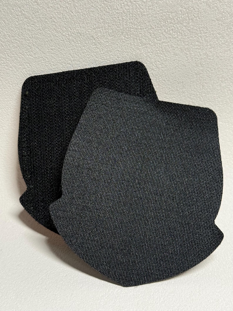 May include: Two black fabric patches with a textured surface. The patches are in a shield shape with rounded corners and a notched bottom. One patch is slightly darker than the other.