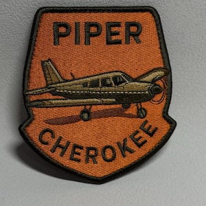 May include: Embroidered patch featuring a Piper Cherokee airplane in a shield shape. The patch has an orange background with the words "PIPER" and "CHEROKEE" in black lettering. The airplane is detailed in shades of brown and black.