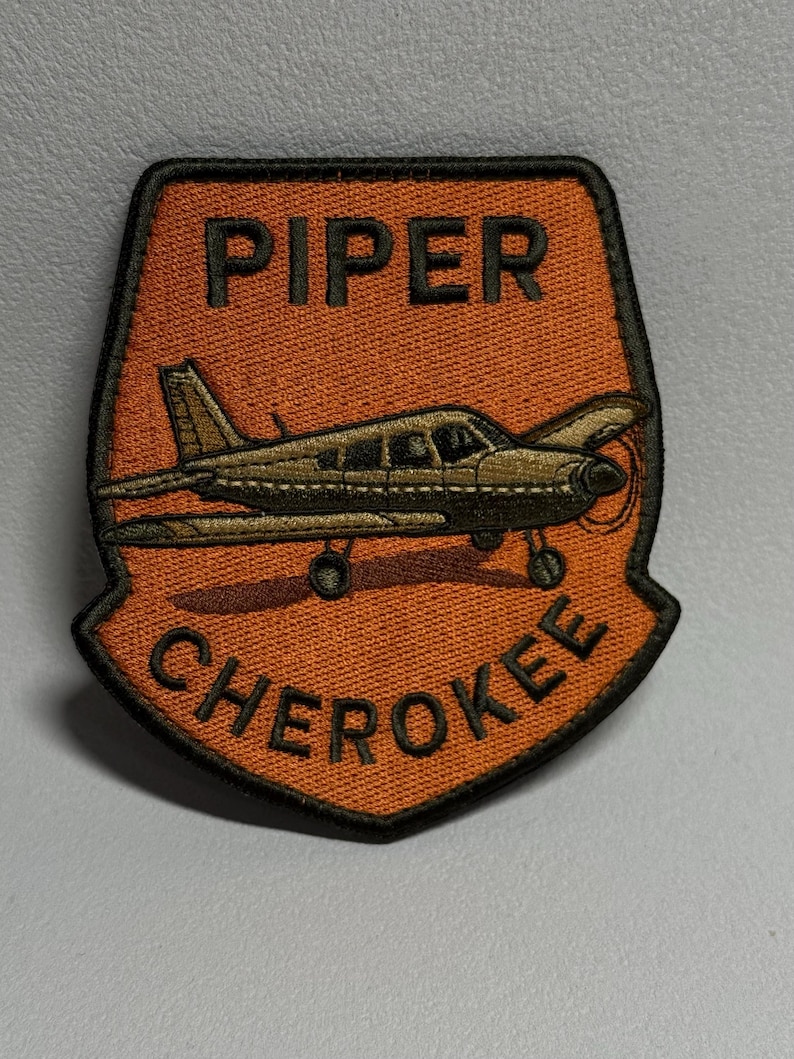 May include: Embroidered patch featuring a Piper Cherokee airplane in a shield shape. The patch has an orange background with the words "PIPER" and "CHEROKEE" in black lettering. The airplane is detailed in shades of brown and black.