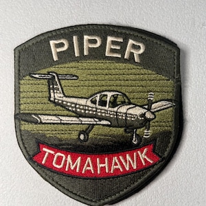 May include: Embroidered patch featuring a silver airplane with a propeller, set against a green and olive background. The patch has the word "PIPER" at the top and "TOMAHAWK" on a red banner.
