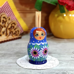 Puede incluir: Un porta palillos de dientes de madera azul y blanco con forma de muñeca rusa con un diseño floral. La muñeca tiene una bufanda roja y blanca con estampado y una cara sonriente con ojos azules. El soporte está sobre un encaje blanco.