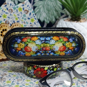 Puede incluir: Un estuche para gafas ovalado de laca negra con un dise&ntilde;o floral. El estuche est&aacute; decorado con una pintura tradicional de arte popular ruso de flores y hojas coloridas. El estuche est&aacute; sentado en un peque&ntilde;o soporte con un dise&ntilde;o floral similar. Un par de gafas de lectura negras y blancas est&aacute;n tiradas sobre una superficie estampada.