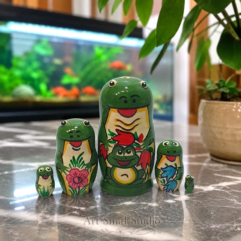 Green frog matryoshka dolls arranged on a marble tabletop in front of a softly blurred aquarium, with five nesting pieces featuring colorful hand-painted floral and frog motifs