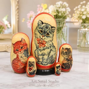 Hand painted Russian nesting dolls with cat portraits displayed on a white marble table in a bright pastel interior with soft blurred flowers, collectible wooden Matryoshka set of 5