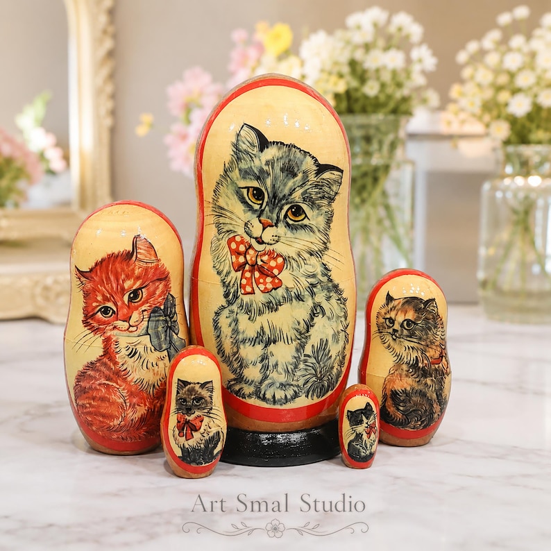 Hand painted Russian nesting dolls with cat portraits displayed on a white marble table in a bright pastel interior with soft blurred flowers, collectible wooden Matryoshka set of 5